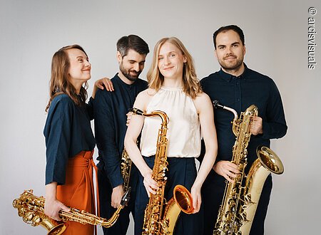 Eine Gruppe von schwarz gekleideten Personen steht zusammen vor einem hellen Hintergrund. Die Personen halten Saxophone in den Händen.