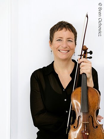 Eine schwarz gekleidete Dame mittleren Alters mit kurzen Haaren hält eine Violine in der Hand und lächelt.