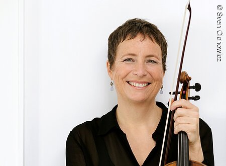 Eine schwarz gekleidete Dame mittleren Alters mit kurzen Haaren hält eine Violine in der Hand und lächelt.