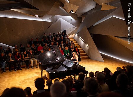 Eine junge Dame im blauen Kleid sitzt an einem Fl¸gel und spielt darauf. Der Raum ist futuristisch mit ineinander geschachtelten riesigen Steinelementen. Im Vorder- und Hintergrund ist unscharf Publikum zu erkennen.
