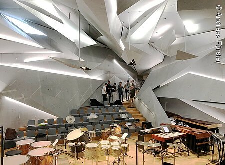 In einem großen, futuristisch gestaltetem Raum sind viele Percussion-Instrumente zu sehen. Im Eingangsbereich stehen junge Menschen mit Percussion-Instrumenten.