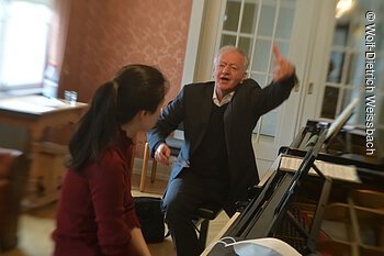 In einem mit Antiquitäten ausgestattetem Raum stehen zwei Flügel, an denen ein älterer Herr und eine junge Dame zusammen sitzen. Die Dame spielt, der Herr gestikuliert.