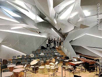 In einem großen, futuristisch gestaltetem Raum sind viele Percussion-Instrumente zu sehen. Im Eingangsbereich stehen junge Menschen mit Percussion-Instrumenten.