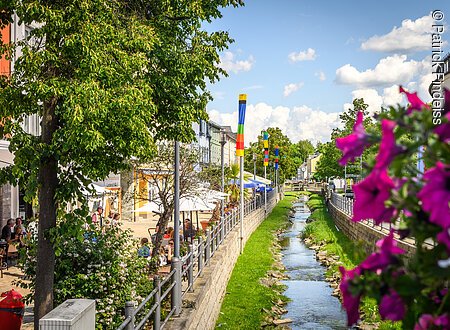 Stadtansicht mit kleinem Fluss, bunten Fahnen, Bäumen und Menschen in einem Café.