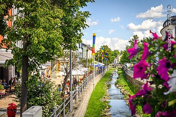 Stadtansicht mit kleinem Fluss, bunten Fahnen, Bäumen und Menschen in einem Café.