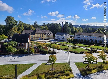 Schlossterrassen (Bad Alexandersbad, Fichtelgebirge) Historisches Gebäude mit gelber Fassade, umgeben von Bäumen und Gärten unter blauem Himmel.