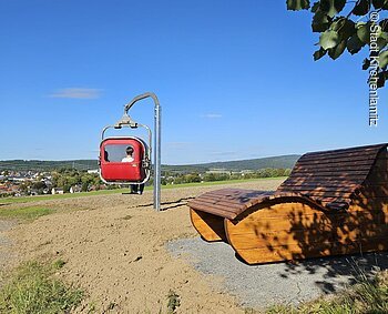 Panorama-Gondel (Kirchenlamitz, Fichtelgebirge) Rote Gondel an einem Mast und eine Holzbank auf einem Hügel mit Blick auf eine Stadt.