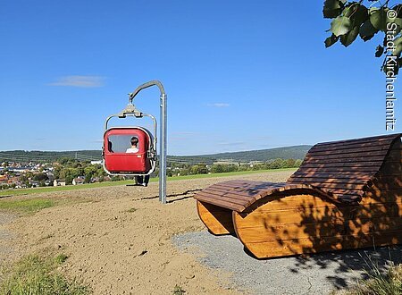 Panorama-Gondel (Kirchenlamitz, Fichtelgebirge) Rote Gondel an einem Mast und eine Holzbank auf einem Hügel mit Blick auf eine Stadt.