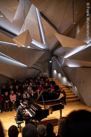 Eine Person sitzt an einem Flügel und spielt darauf. Im Hintergrund ist unscharf Publikum zu erkennen. Der Raum ist futuristisch mit ineinander geschachtelten riesigen Steinelementen.