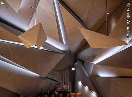 Eine Person sitzt an einem Flügel und spielt darauf. Im Hintergrund ist unscharf Publikum zu erkennen. Der Raum ist futuristisch mit ineinander geschachtelten riesigen Steinelementen.