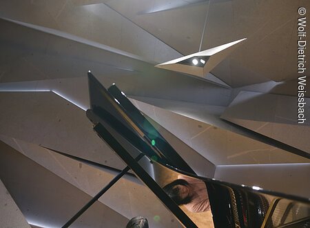 Eine Dame spielt an einem Flügel in einem modern gestalteten Raum, der aus Steinelementen besteht und geheimnisvoll beleuchtet ist..Der Blick geht durch den aufgestellten Flügel.