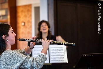 Eine junge Dame bläst in eine Oboe, neben ihr eine Dame, die mit ihr spricht und gestikuliert.