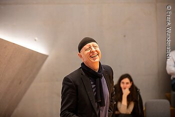 Ein Herr mit einer schwarzen Mütze steht vor einer Betonwand und lacht. Im Hintergrund ist unscharf eine sitzende junge Frau zu sehen.