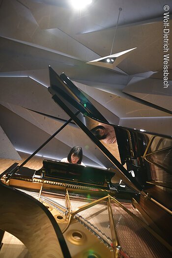 Eine Dame spielt an einem Flügel in einem modern gestalteten Raum, der aus Steinelementen besteht und geheimnisvoll beleuchtet ist..Der Blick geht durch den aufgestellten Flügel.