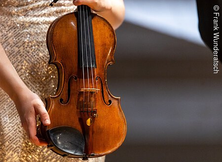 Eine Hand  hält eine Violine, hochkant. Der Hals des Instruments ist nicht zu sehen.