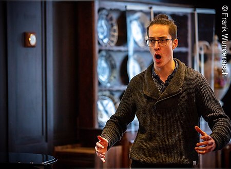 Ein junger Mann singt in einem holzvertäfelten Raum. Im Hintergrund ist eine antike Porzellanvitrine zu sehen.