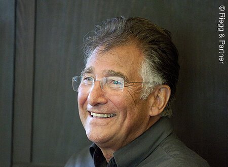 Ein älterer Mann mit rahmenloser rechteckiger Brille sitzt vor einer Holzwand. Er schaut an der Kamera vorbei und lacht.