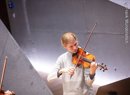 Eine junge Frau im grauen Rollkragenpullover spielt Geige. Im Raumhintergrund sind geometrische Formen mit effektvoller Beleuchtung zu erkennen. Am linken Bildrand sind Teile einer Geige und eines Bogens zu sehen.