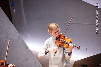 Eine junge Frau im grauen Rollkragenpullover spielt Geige. Im Raumhintergrund sind geometrische Formen mit effektvoller Beleuchtung zu erkennen. Am linken Bildrand sind Teile einer Geige und eines Bogens zu sehen.