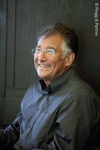 Ein älterer Mann mit rahmenloser rechteckiger Brille sitzt vor einer Holzwand. Er schaut an der Kamera vorbei und lacht.