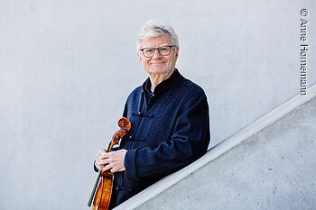 Ein älterer Mann steht hinter einer schräg verlaufenden, halbhohen Betonwand. Er stützt sich mit einem Arm darauf ab und hält eine Viola vor dem Oberkörper.