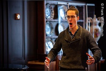 Ein junger Mann singt in einem holzvertäfelten Raum. Im Hintergrund ist eine antike Porzellanvitrine zu sehen.