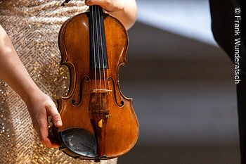 Eine Hand  hält eine Violine, hochkant. Der Hals des Instruments ist nicht zu sehen.